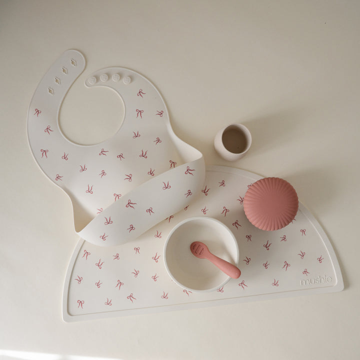 Baby bib, mat, and cup set with pink accents on a light background