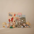 A wooden farm animal puzzle displayed upright on a flat surface, surrounded by an arrangement of various baby toys, teethers, and rattles in neutral and pastel tones.