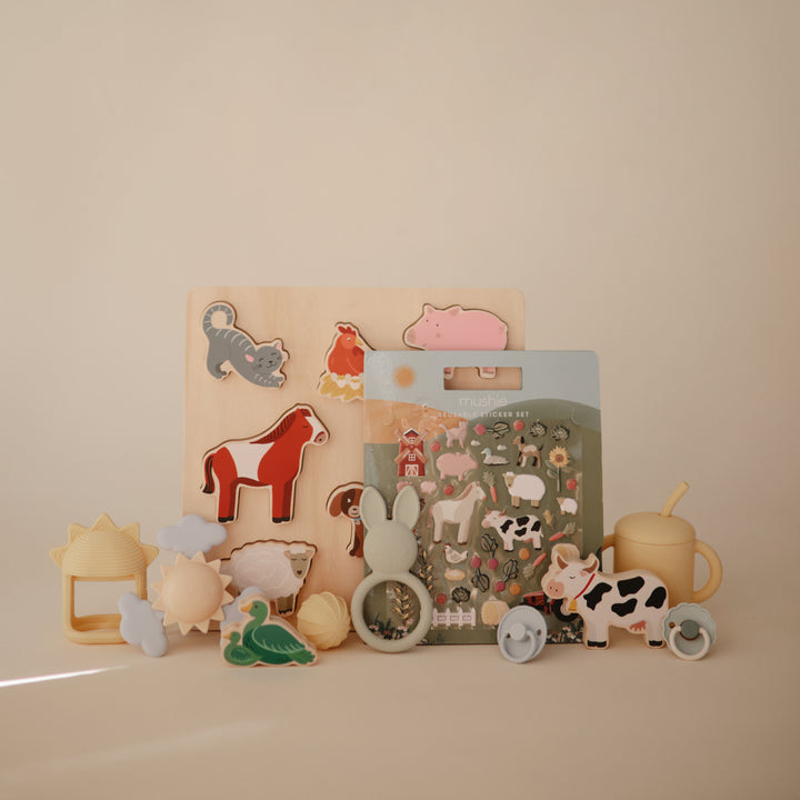 A wooden farm animal puzzle displayed upright on a flat surface, surrounded by an arrangement of various baby toys, teethers, and rattles in neutral and pastel tones.