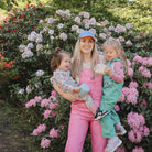 Woman in pink overalls holding two children in a garden with pink flowers