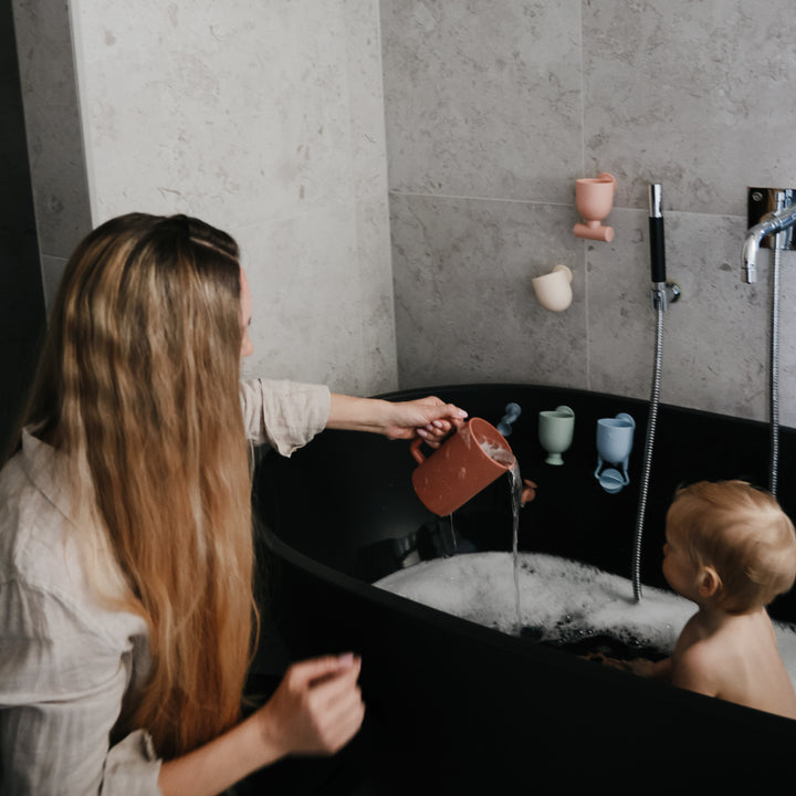 Woman pouring water from a pitcher into a bathtub with a child inside, in a bathroom setting.