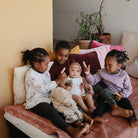 Four children sitting together on a bed with pillows and plants in the background