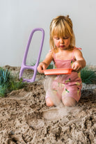 Child playing in sand with a pink shovel and orange pan on a sandy surface.