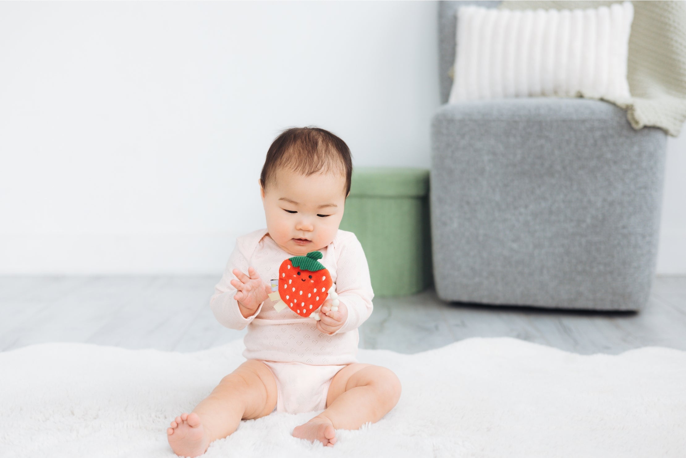 Baby sitting on a white blanket holding a toy strawberry in a minimalistic room.