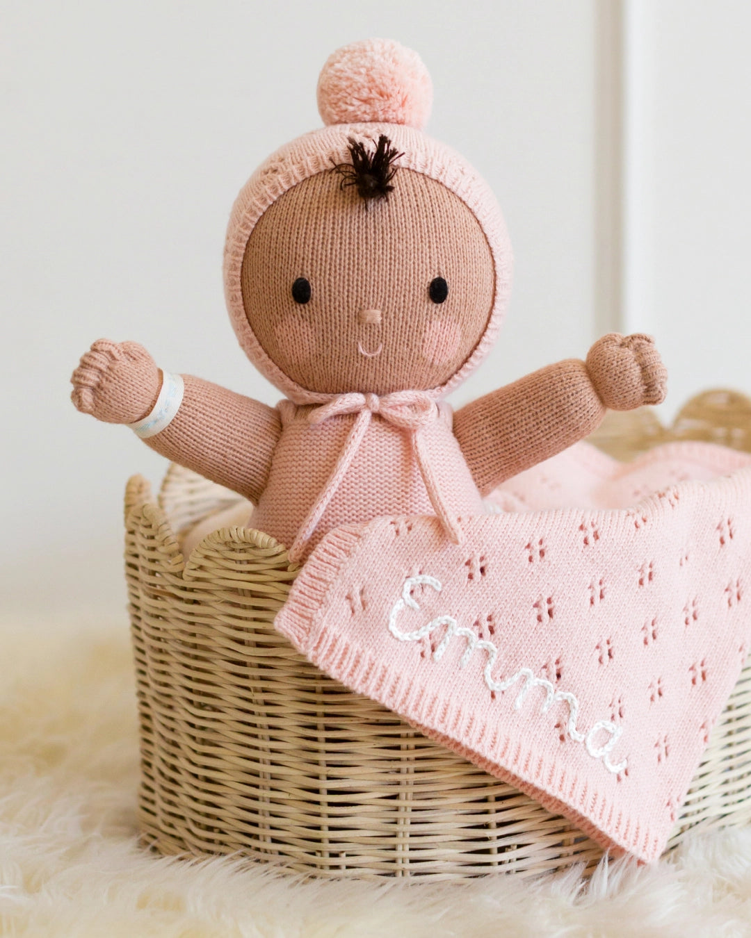 A knitted doll in a pink bonnet with a pom-pom and bow, posed in a wicker basket with a pink knitted blanket that says 'Emma' in white script.
