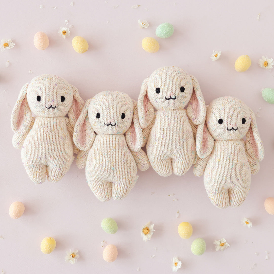 Four cream-colored knit bunny toys on a light pink background with small pastel eggs and flowers.