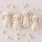 Four cream-colored knit bunny toys on a light pink background with small pastel eggs and flowers.