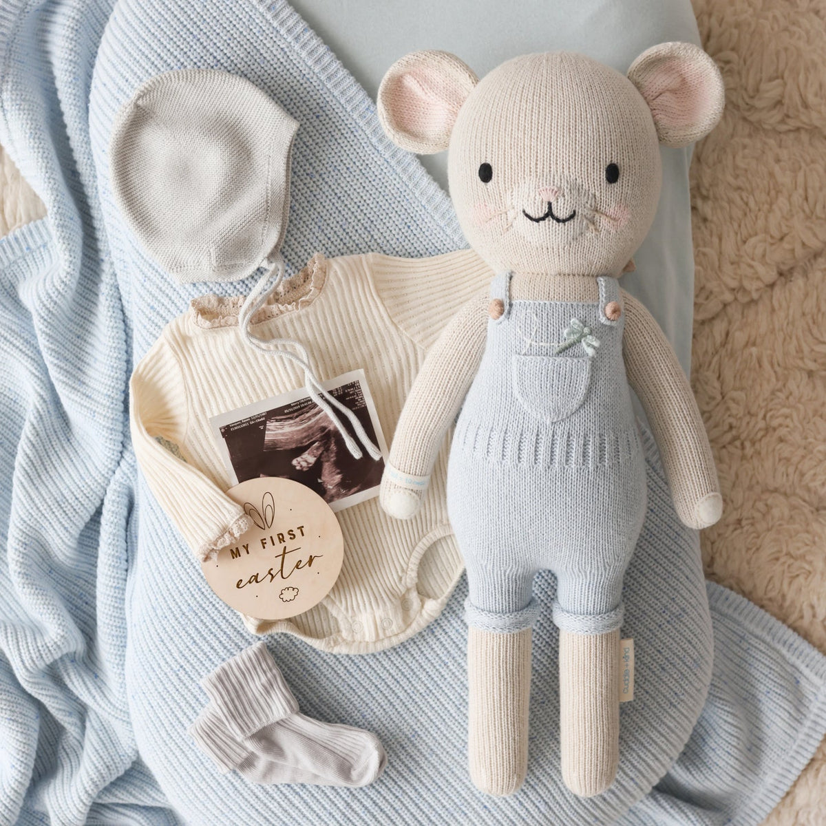 Knitted toy mouse in overalls surrounded by baby items on a textured blanket
