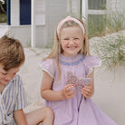 A young girl with blonde hair sits on a sandy beach next to a boy, wearing a lavender dress with blue embroidery and holding a butterfly-shaped accessory. A white beach house and dune grass are in the background.