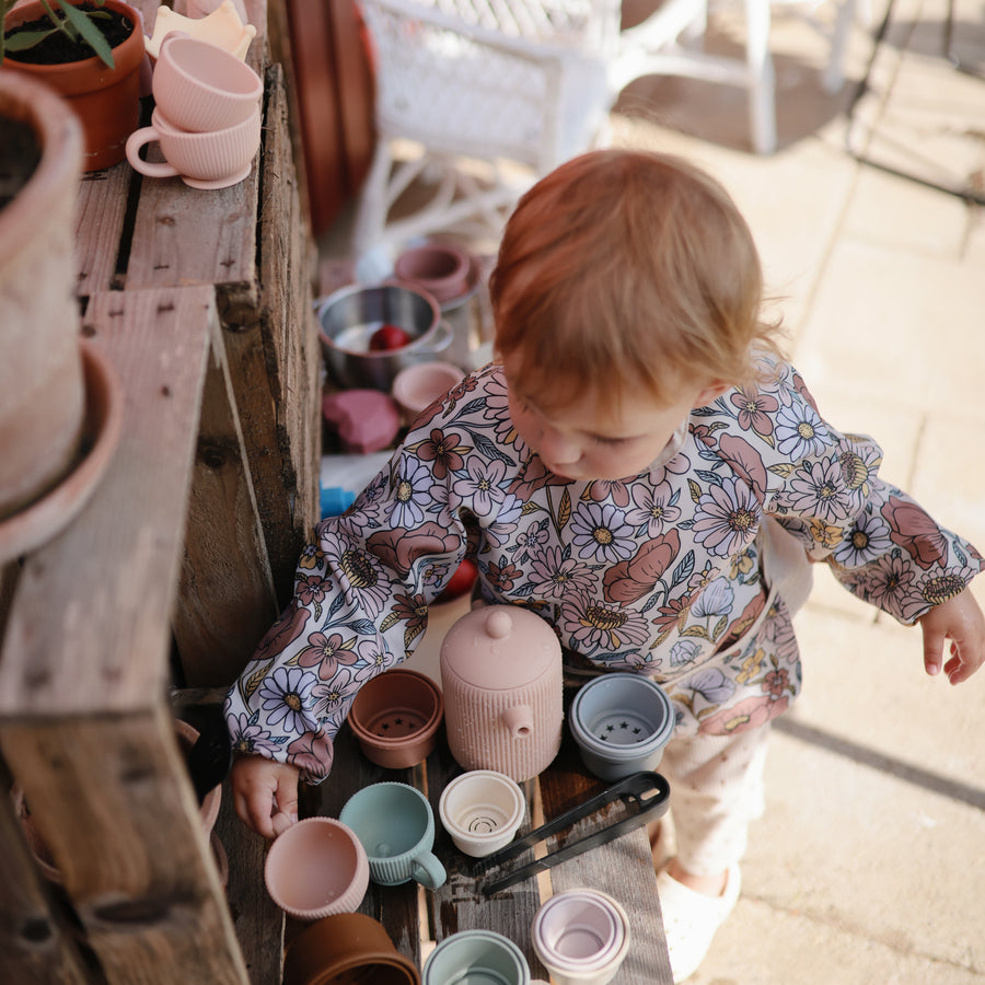 A young child wearing a floral print top reaches toward a wooden surface arranged with a silicone tea set and small pots outdoors.