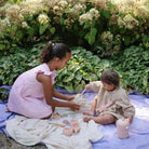 Two young girls playing with toys on a blanket outdoors surrounded by greenery.