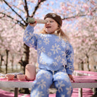 A young child wearing a matching blue floral outfit and a brown beanie, sitting at an outdoor table with cherry blossoms in bloom, holding up a Mushie kaleidoscope to one eye.