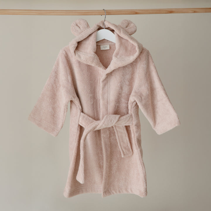 Pink children's robe with bear ears on a hanger against a beige wall.