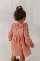 A toddler girl seen from behind wearing a dusty pink long-sleeve cotton gauze dress with a ruffled neckline and small embroidered floral motifs, standing against a white studio background.