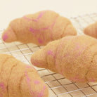 Felted wool croissants in a light tan color with pink markings, resting on a metal cooling rack.