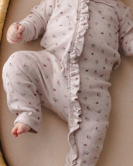 A close-up of a baby wearing a light pink long-sleeve footless sleeper with a delicate multicolor floral print and ruffle trim down the front, lying on a beige blanket.
