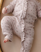 A close-up of a baby wearing a light pink long-sleeve footless sleeper with a delicate multicolor floral print and ruffle trim down the front, lying on a beige blanket.