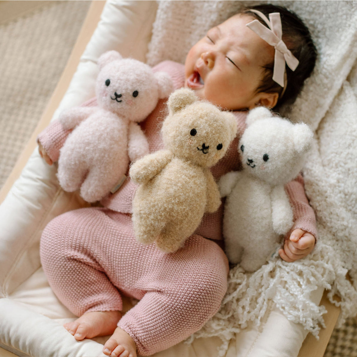 Baby in pink outfit holding three teddy bears on a soft surface