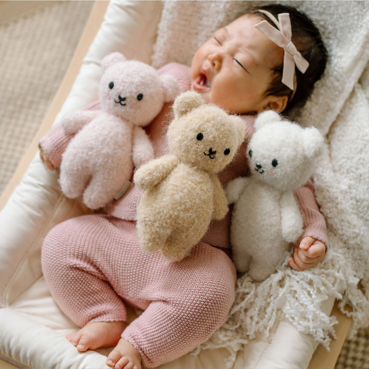 Baby in pink outfit holding three teddy bears on a soft surface