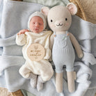 Newborn baby in a cream outfit with 'My First Easter' sign, lying next to a plush toy on a textured blanket.