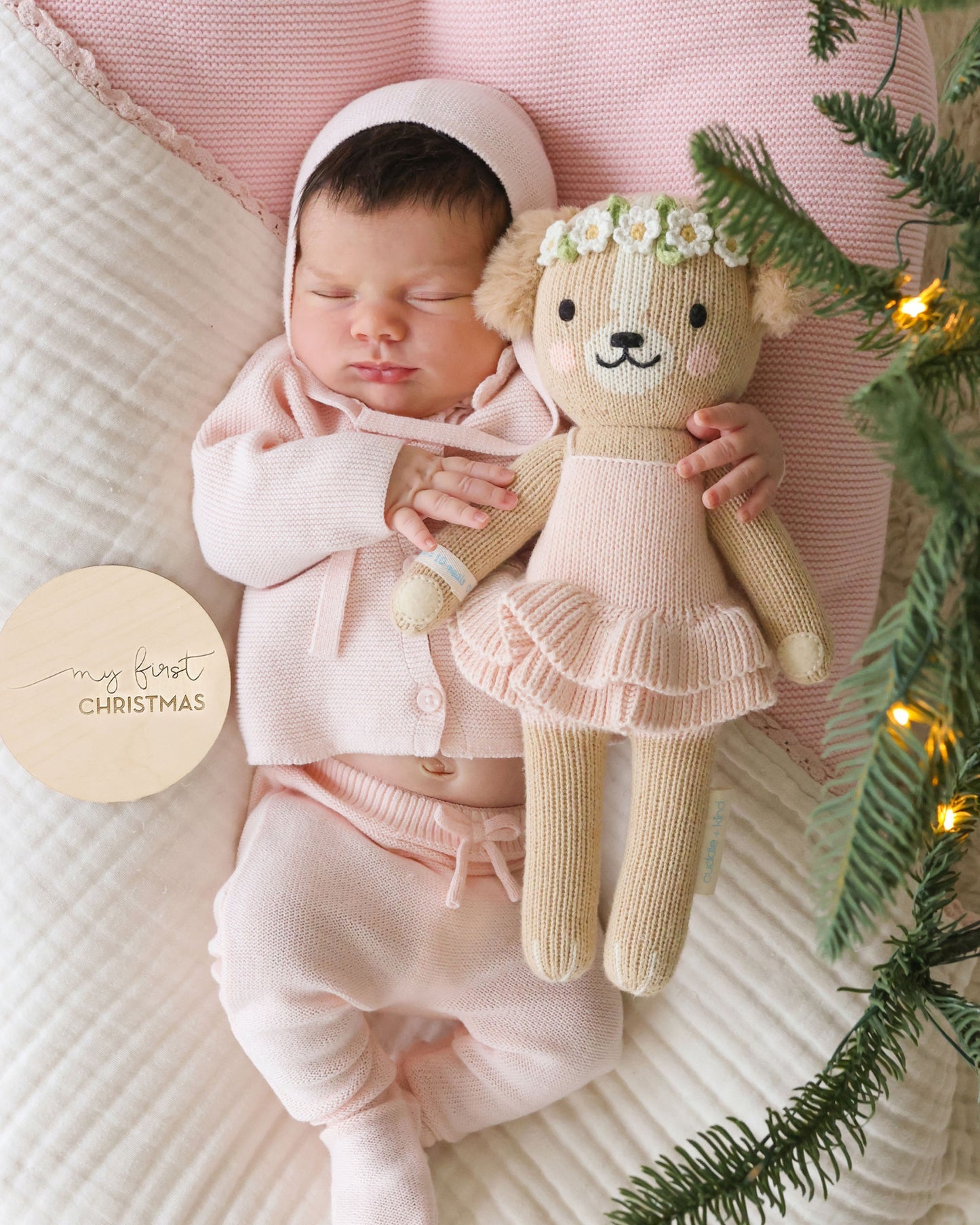 A sleeping newborn baby wearing a soft pink knit bonnet, cardigan, and footed pants, cuddling a knitted teddy bear doll with a flower crown. The baby is lying on a textured cream quilt with a pink pillow, and evergreen branches with warm string lights frame the scene.