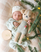 Newborn baby in a green outfit holding a teddy bear next to Christmas tree branches.