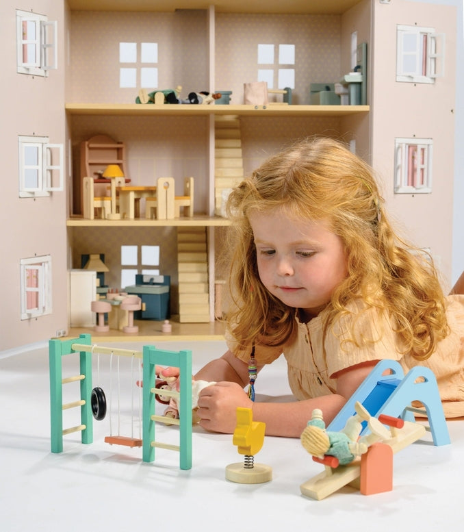 A young child playing on the floor with a large wooden dollhouse, accompanied by a wooden playground set that includes a slide, see-saw, climbing frame, swing, tire, and a bouncy bunny toy.