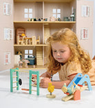 A young child playing on the floor with a large wooden dollhouse, accompanied by a wooden playground set that includes a slide, see-saw, climbing frame, swing, tire, and a bouncy bunny toy.