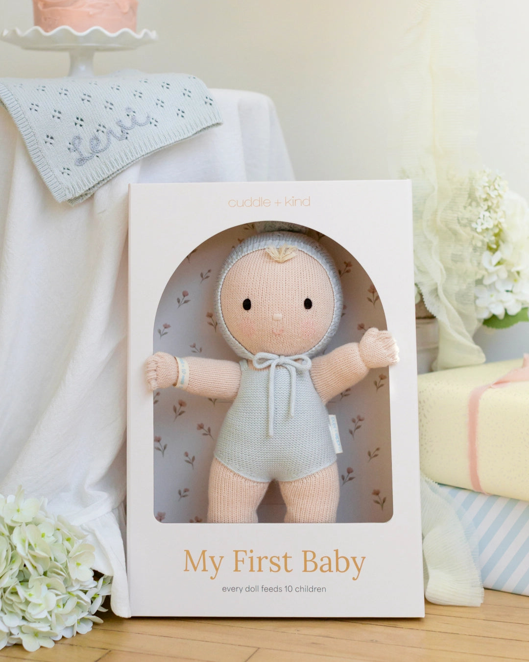 A knitted baby doll with a pale peach face and limbs, wearing a light blue knit bonnet with a pom-pom and a matching blue knitted romper.