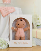Baby doll in a biege outfit in a box labeled 'My First Baby' with a pink blanket and flowers in the background.