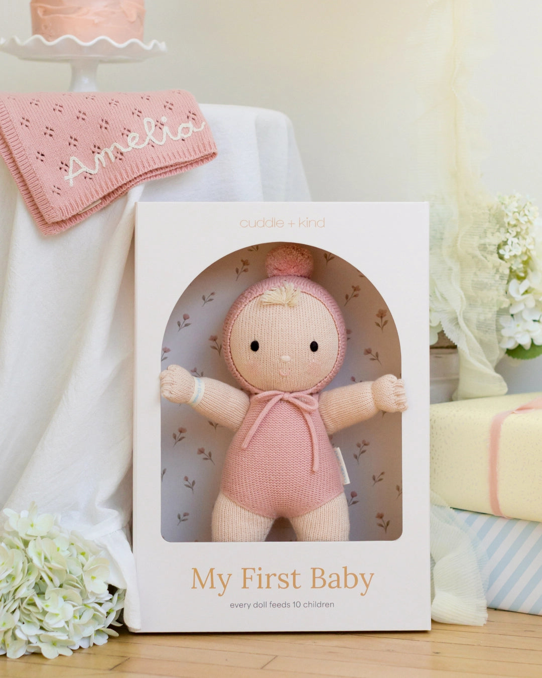 A knitted baby doll in a white gift box with the text 'My First Baby' and 'every 2nd baby needs 10 children.' The doll is pale pink and cream with a pink bonnet and bow.