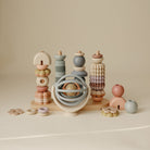 A collection of Mushie baby toys, including a Suction Spinner Ball, a wooden stacking tower, interlocking rings, and various wooden and silicone play pieces arranged on a neutral surface.