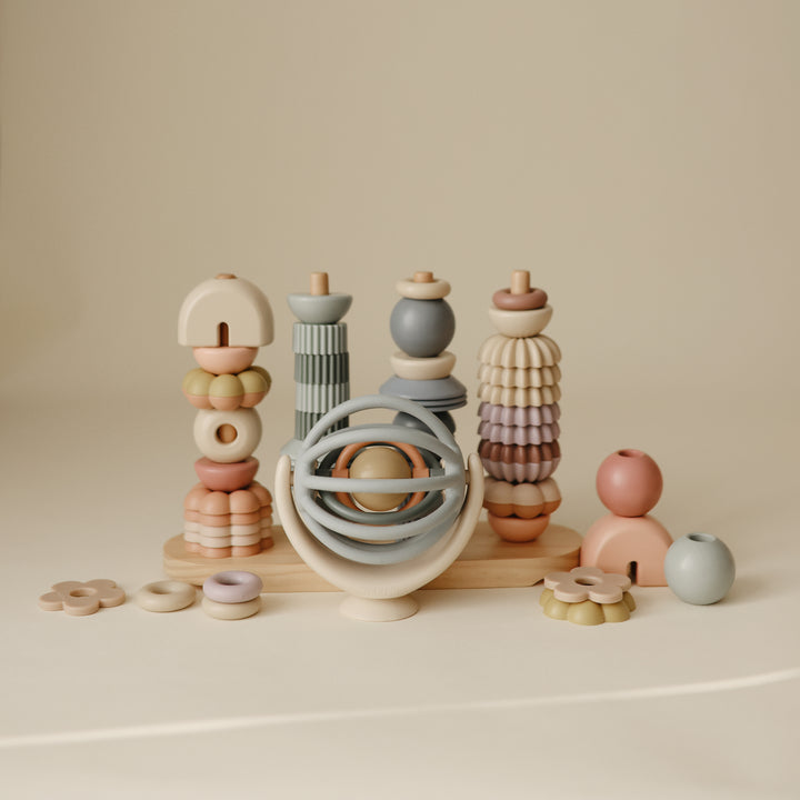 A collection of Mushie baby toys, including a Suction Spinner Ball, a wooden stacking tower, interlocking rings, and various wooden and silicone play pieces arranged on a neutral surface.