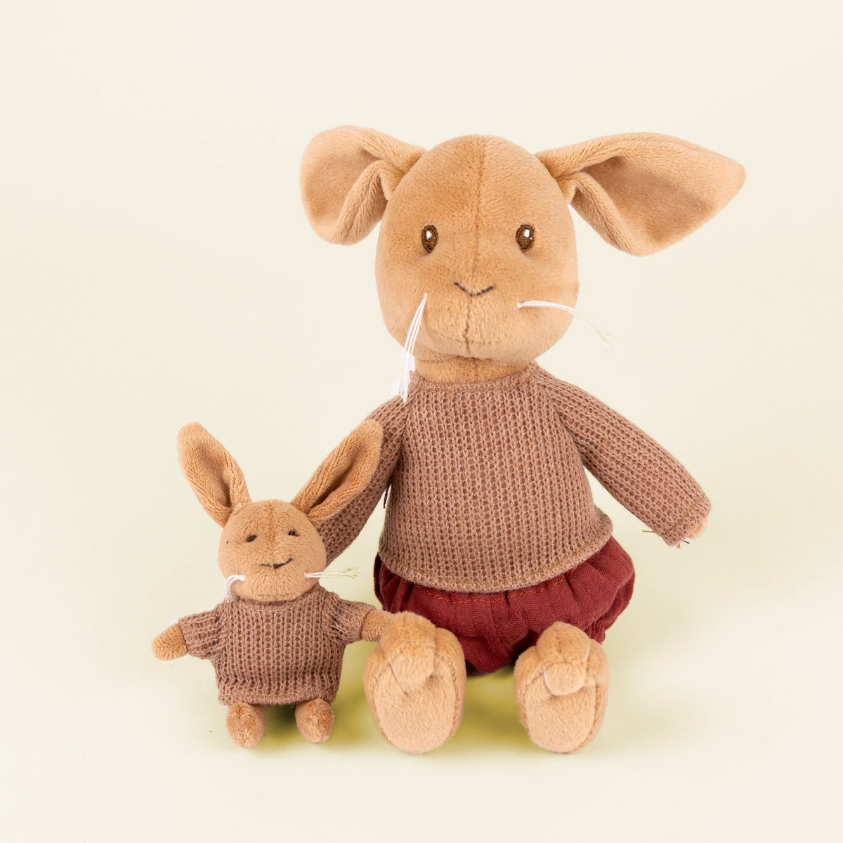 Two plush stuffed rabbits posed together on a plain light cream background. The larger rabbit wears a brown knit sweater and burgundy pants, while the smaller rabbit wears a matching brown knit sweater.