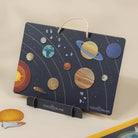 A children's wooden solar system toy set by Coco Village, featuring a dark blue board with 3D planet cutouts, white orbital lines, and a white hanging loop, displayed on a stand with a few loose space-themed pieces nearby.