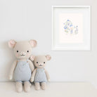 Two stuffed animals in light blue outfits sitting on a white surface with a framed artwork in the background.
