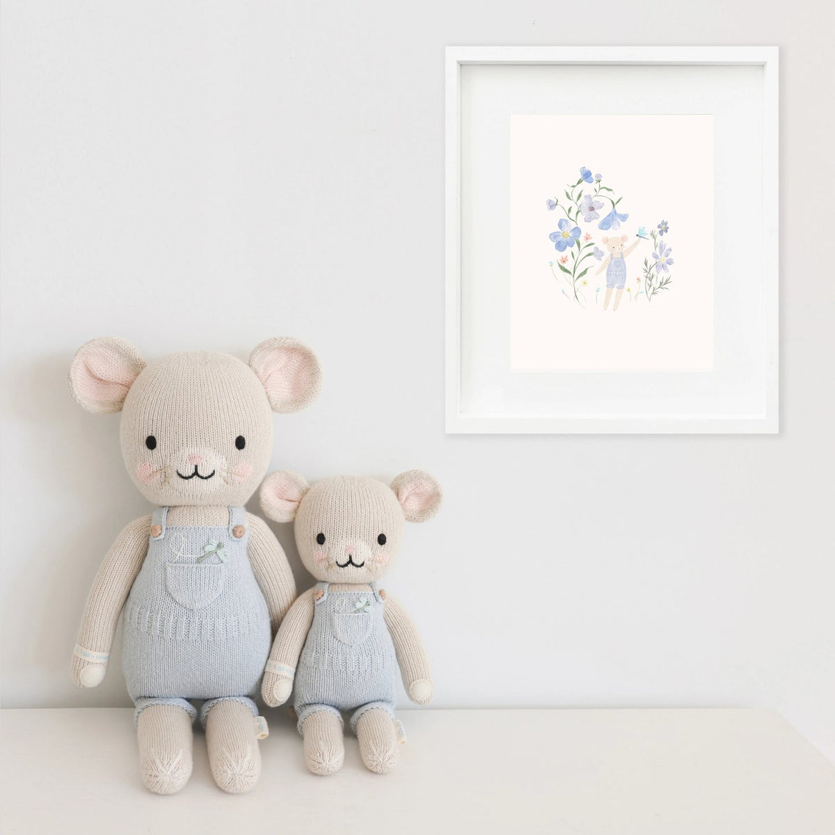 Two stuffed animals in light blue outfits sitting on a white surface with a framed artwork in the background.