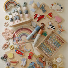 A flat lay arrangement on a white background featuring two Mushie kaleidoscopes surrounded by wooden toys, silicone stackers, animal figurines, an abacus, and space-themed pieces.