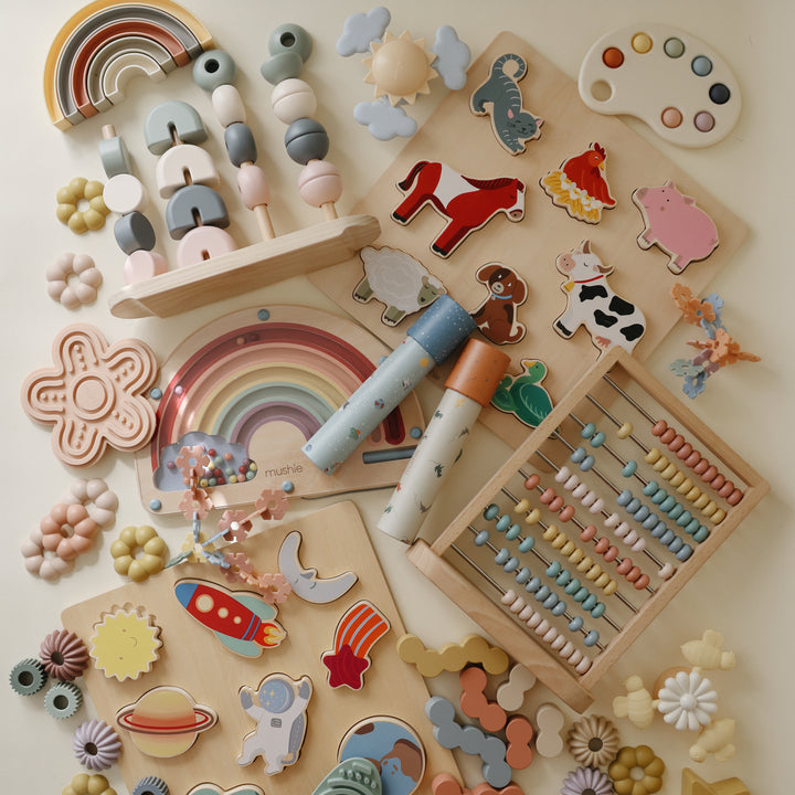 A flat lay arrangement on a white background featuring two Mushie kaleidoscopes surrounded by wooden toys, silicone stackers, animal figurines, an abacus, and space-themed pieces.