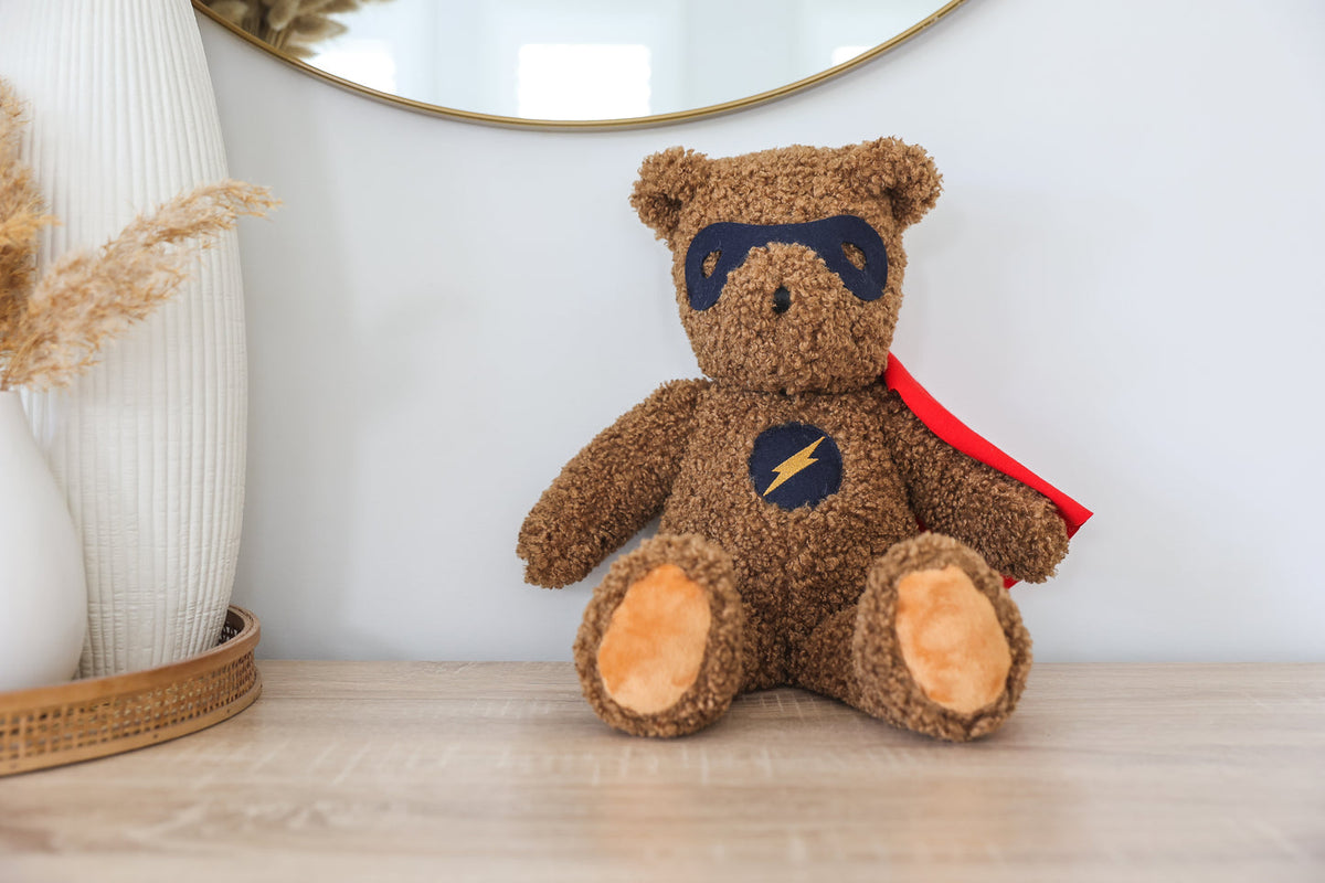 Brown teddy bear with superhero cape and mask on a wooden surface.