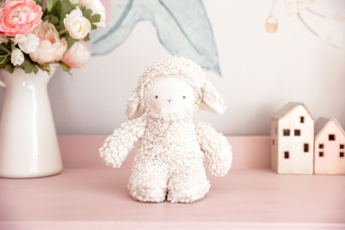 A white textured plush sheep toy sits centered on a pink surface, with a vase of pink flowers on the left and two small house-shaped ornaments on the right against a soft pastel background.