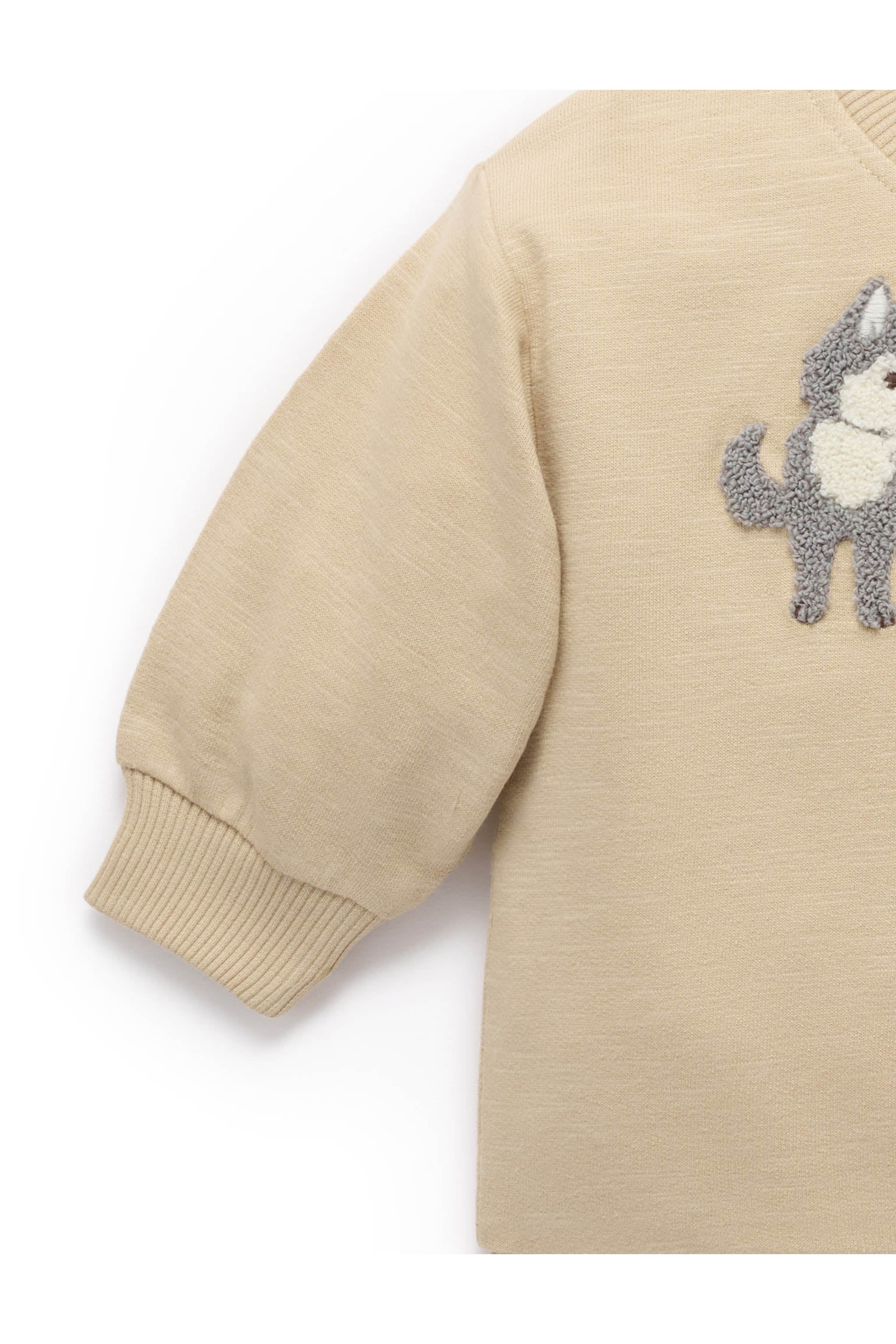 Close-up of a beige long-sleeve knit top with a gray and white dog patch on the chest, showing the ribbed cuff and part of the body against a white background.
