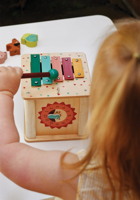 A child's hand holding a red mallet to play the colorful xylophone on top of the wooden activity cube by Mentari, viewed from over the child's shoulder.