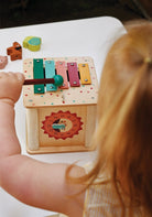 A child's hand holding a red mallet to play the colorful xylophone on top of the wooden activity cube by Mentari, viewed from over the child's shoulder.