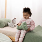 A young child wearing pink and white tie-dye pajamas sits on a bed holding a green plush hippo toy, with a white plush unicorn toy on the bed in the background.