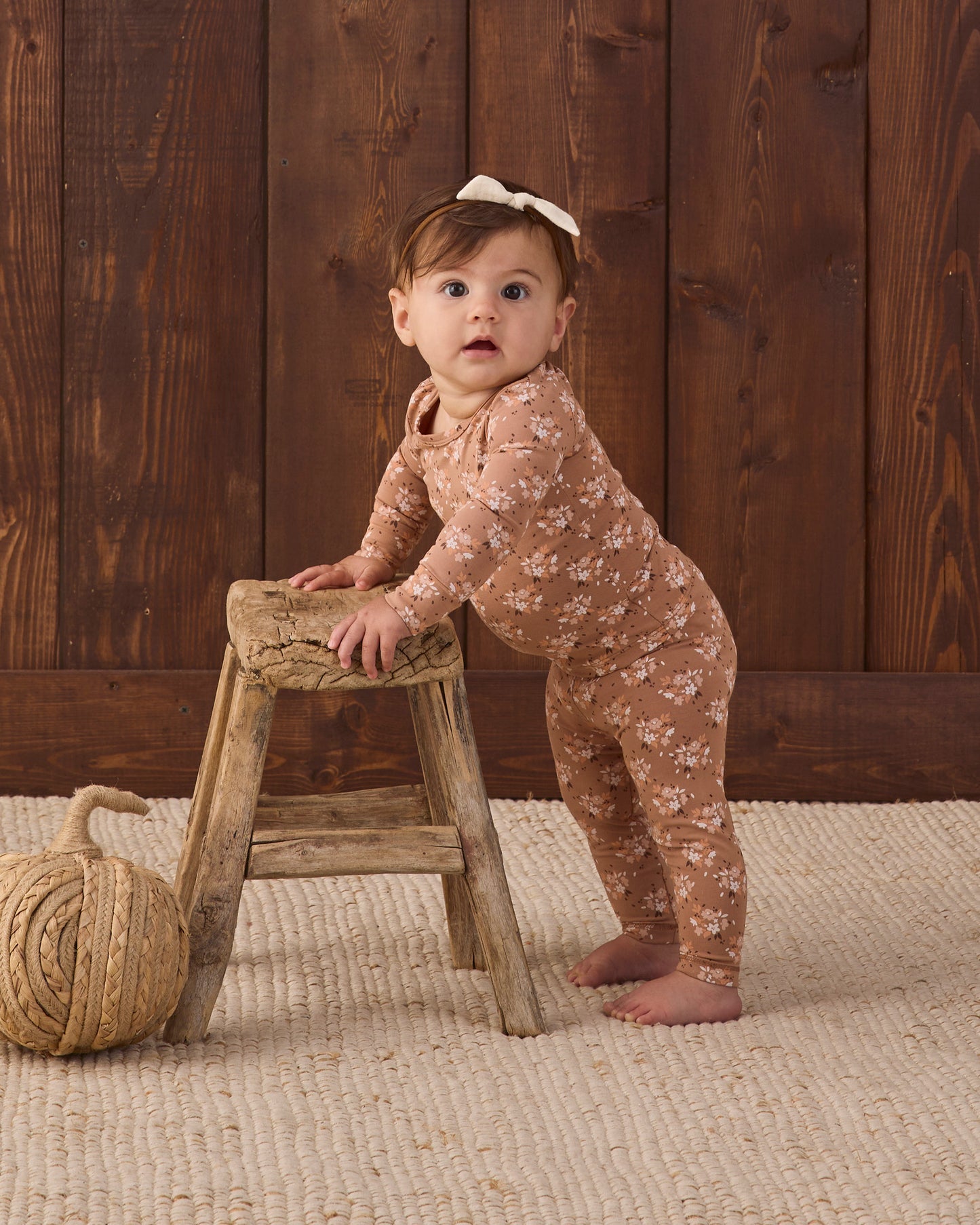 A baby girl wearing a brown long-sleeve floral jumpsuit and a white bow headband stands on a woven rug, leaning on a rustic wooden stool in front of a dark wood plank wall.