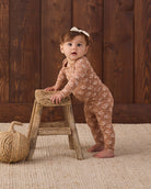A baby girl wearing a brown long-sleeve floral jumpsuit and a white bow headband stands on a woven rug, leaning on a rustic wooden stool in front of a dark wood plank wall.