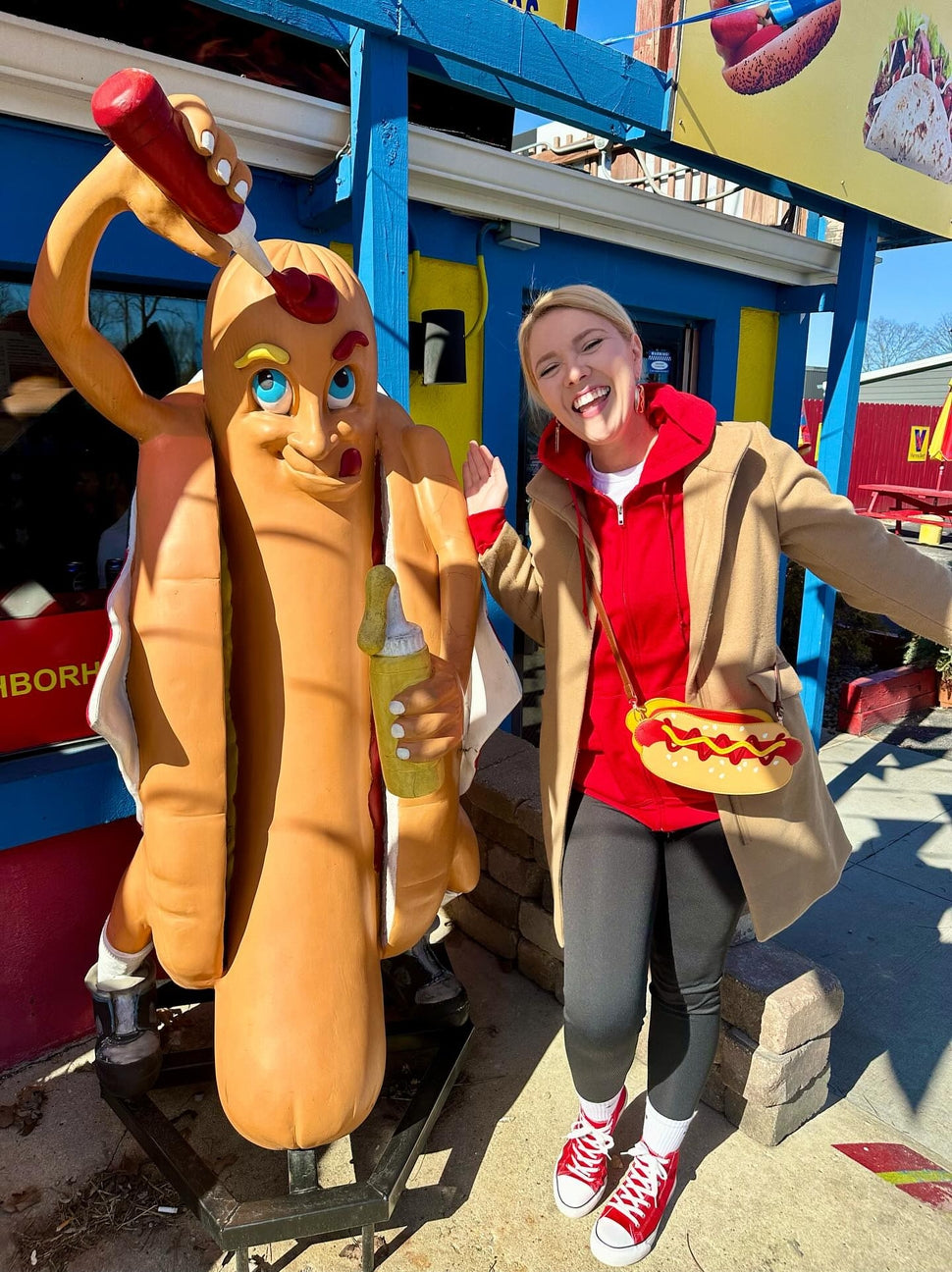 Person in a hot dog costume standing next to a hot dog statue in front of a colorful building.