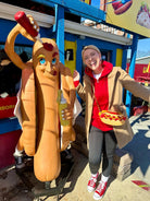 Person in a hot dog costume standing next to a hot dog statue in front of a colorful building.