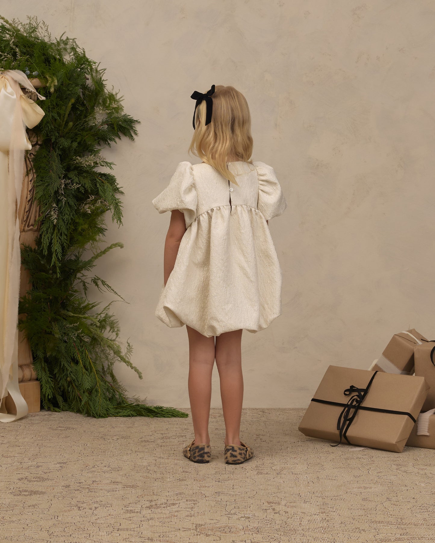 A young girl with blonde hair tied back with a black bow is shown from behind wearing a short cream-colored puff-sleeve dress. She is standing in a styled studio setting with a textured beige wall, a green foliage arrangement on the left, and wrapped gift boxes on the floor to the right.